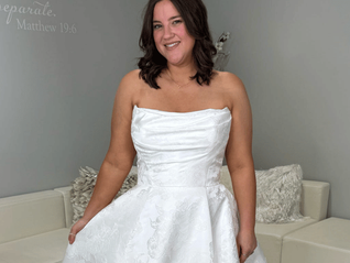 Smiling bride in a white strapless wedding dress stands by a beige couch; wall text reads Matthew 19:6.