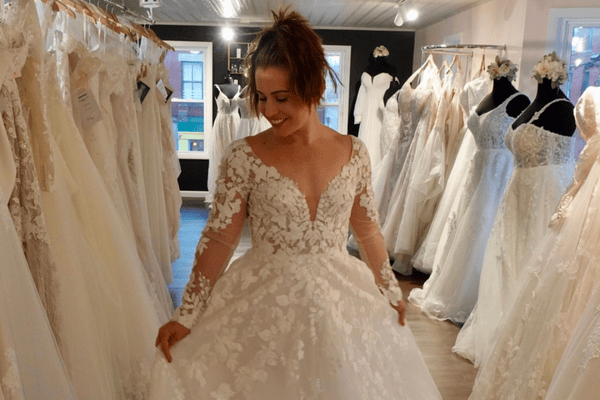 A smiling woman in a lace wedding dress twirls in a bridal shop filled with white dresses, wondering how long to order wedding dress in advance to ensure the perfect fit for her big day.