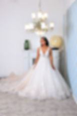 A woman in a white wedding dress stands under a chandelier in a stylish room with light blue wallpaper and a green vase on the mantle.