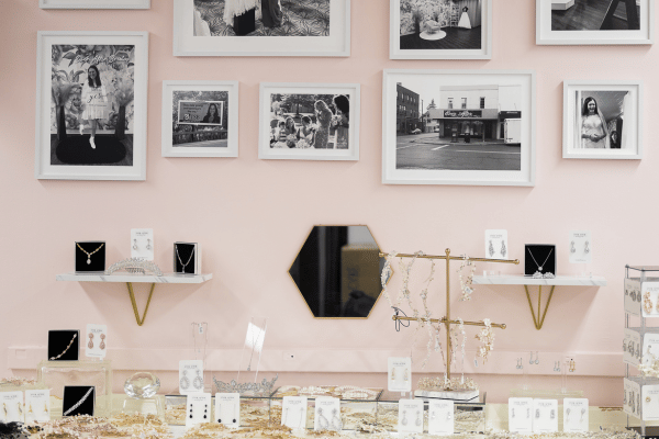 Jewelry display on a pink wall with framed photos above, gold and black necklaces and earrings on shelves and counters.