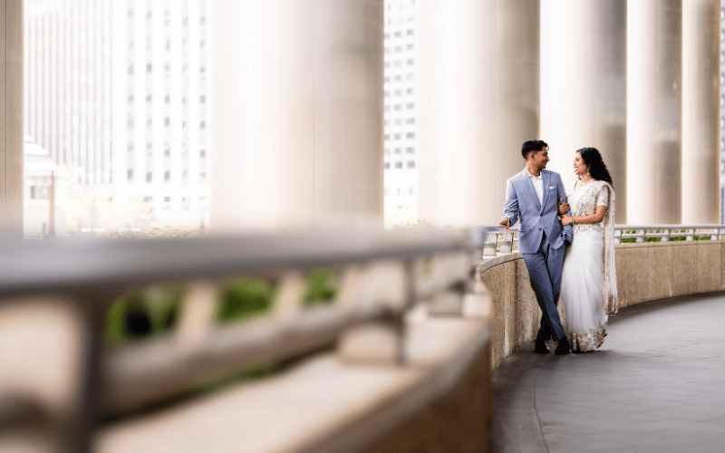 Smiling couple in wedding attire walk arm in arm on a curved balcony with tall columns and city buildings in the background, wedding photo tips