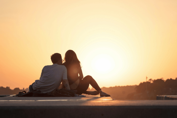 Couple sitting close together on a dock at sunset, silhouetted against a glowing orange sky, sharing a quiet romantic moment.