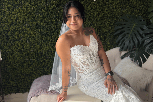 Smiling bride in a white lace wedding dress sits on a couch before a green plant wall, with a large leaf and soft pillows.