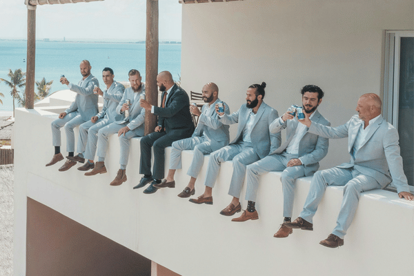 Seven men in light blue suits toast with drinks while sitting on a white balcony overlooking the sea, relaxed and celebratory.