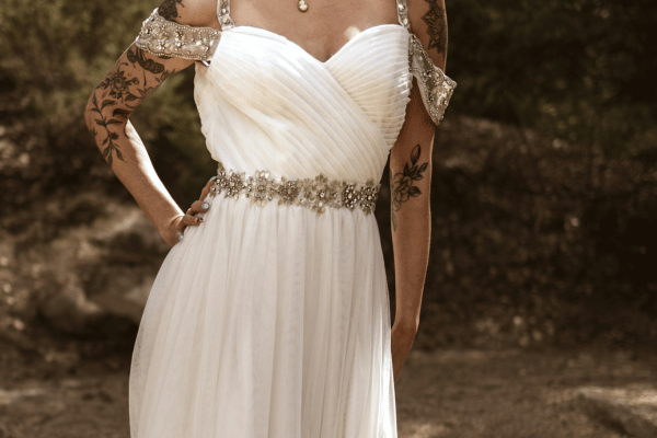 Woman in a white beaded strapless wedding dress with floral tattoos stands outdoors against a blurred rocky background.