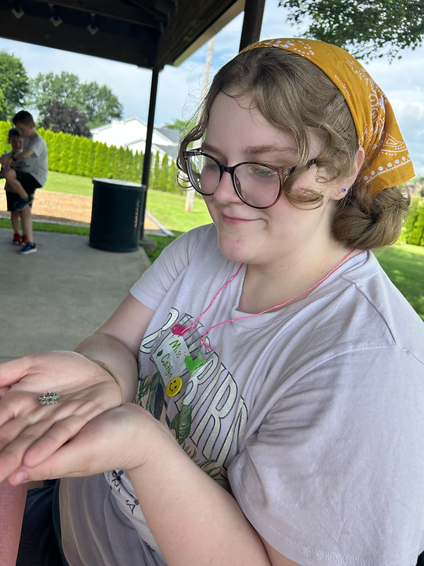 Young woman smiling, holding a caterpillar, wearing glasses and a bandana outdoors.