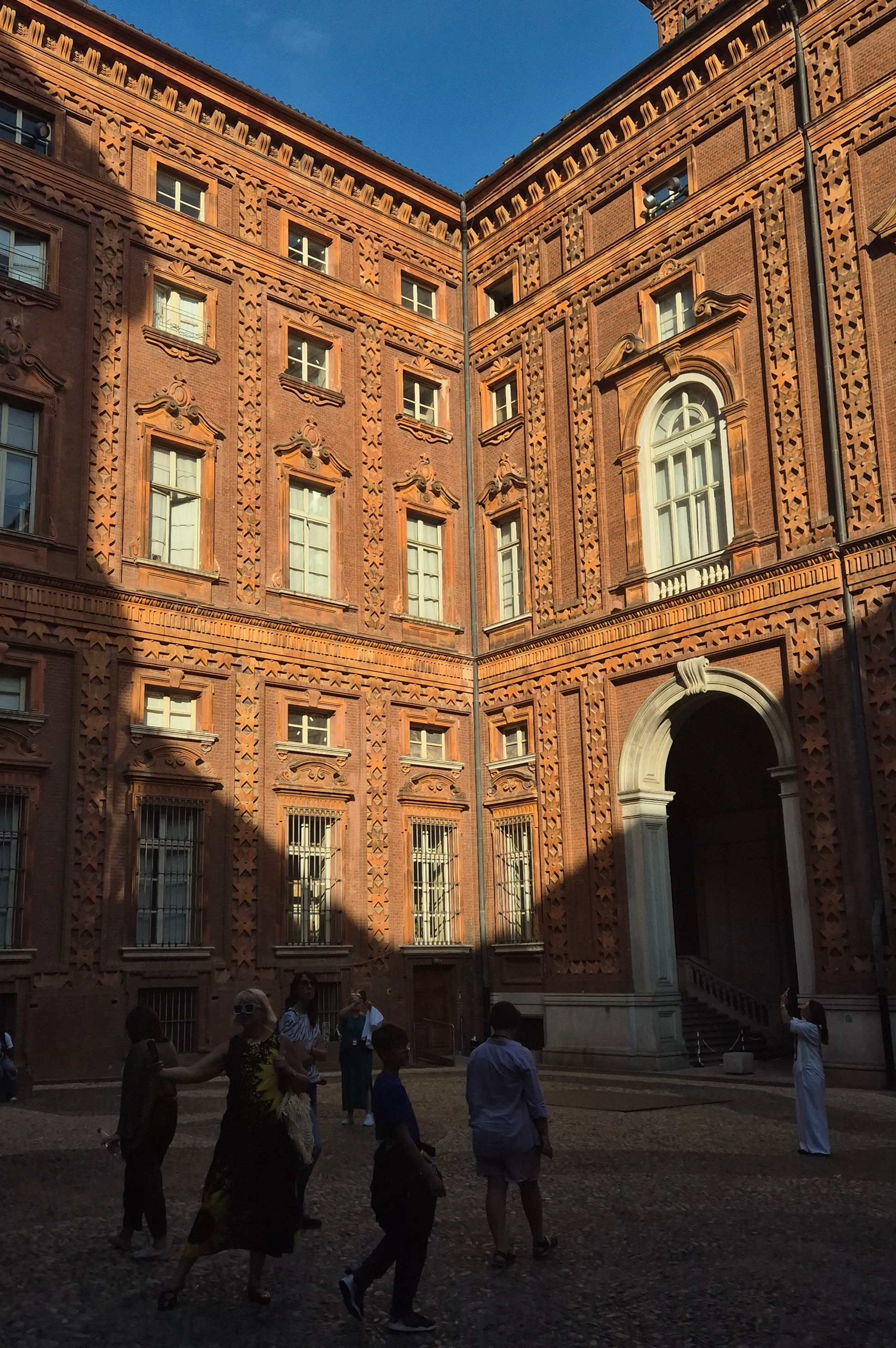 Tourists stand in a sunlit brick courtyard with ornate historic walls and arched doorway under a clear blue sky.
