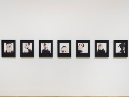 A series of nine framed black-and-white portraits displayed on a plain wall, showing various poses and expressions, conveying introspection.