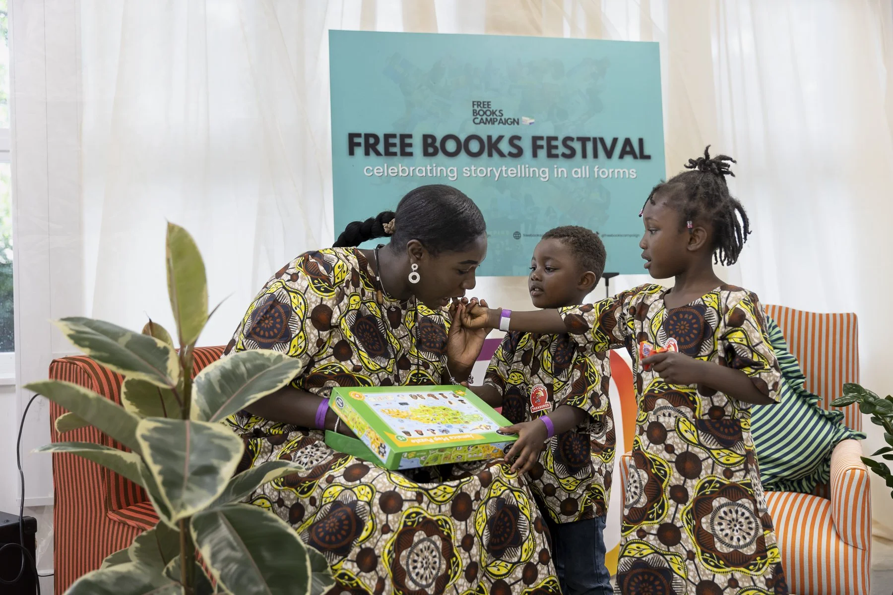 Woman and two children in matching patterned outfits at Free Books Festival, sharing a board game in a bright booth.