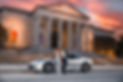 Couple dressed in formal wear in front of car in front of building.
