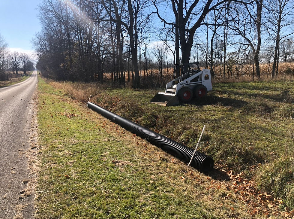 A Bobcat loader near a long black pipe by a rural road. Bare trees and grassy field in background under clear blue sky.