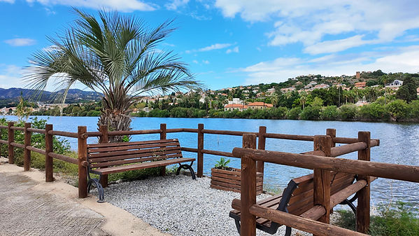 Scenic view of a tranquil lakeside spot in Ganzirri with wooden benches, palm trees, and calm blue waters under a clear sky.