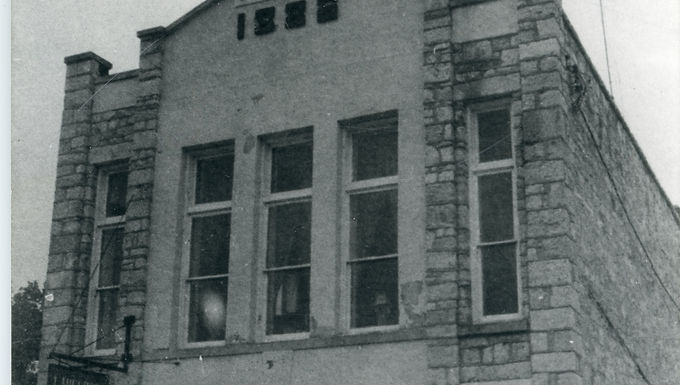 Historic photo of the Old Village Hall in Lanesboro, Minnesota, a stone building dated 1895 with large upper windows, a bell tower, and double garage doors on the ground floor.
