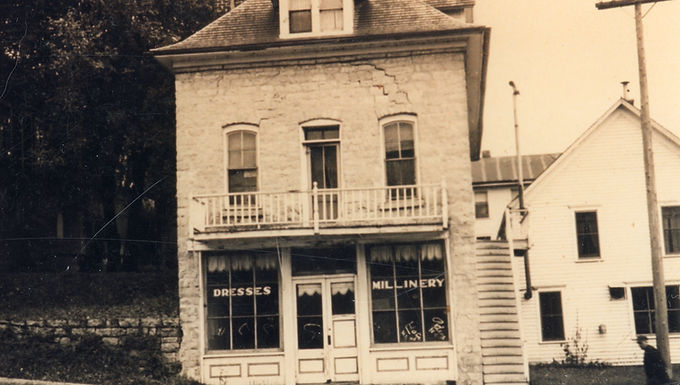 Historic photo of the two-story stone Legion Building with a balcony, displaying shop windows labeled “Dresses” and “Millinery,” and an exterior staircase on the right side.