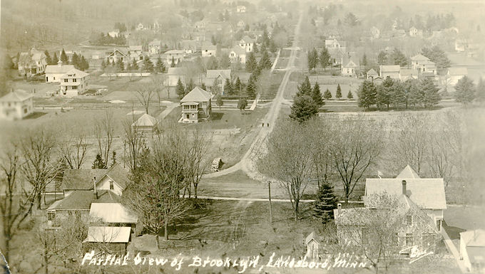 Historic panoramic view of Lanesboro, Minnesota, showing the Brooklyn neighborhood with tree-lined streets, houses, and open yards in a lightly wooded landscape.