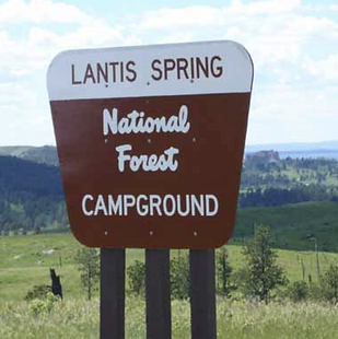 This photo shows the entrance sign for Lantis Spring National Forest Campground in Montana. The brown-and-white sign stands prominently in a grassy landscape with rolling green hills and scattered trees stretching into the distance under a bright sky.