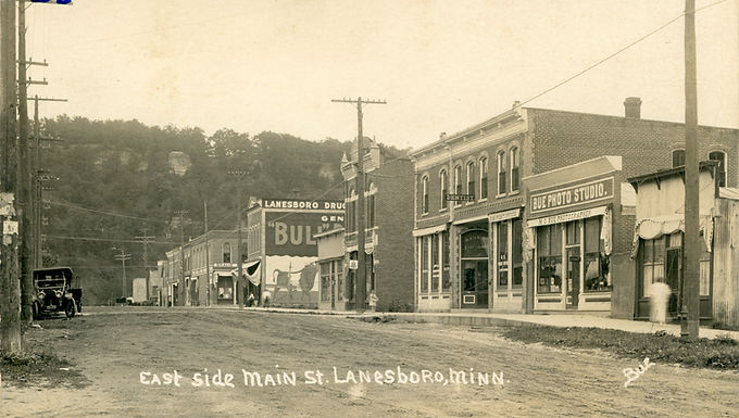 Historic photo of East Main Street in Lanesboro, Minnesota, featuring storefronts including Bue Photo Studio, a dentist’s office, and Lanesboro Drug, with a painted wall advertisement and early automobile on the dirt road.