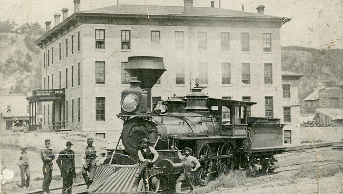 A vintage photograph of a steam locomotive with several men posing beside it, set in front of a large brick building, in the small town of Lanesboro, MN.