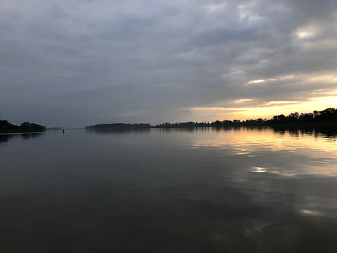 A rower is in the distance on a section of the river, the water is flat, with a sunrise appearing.