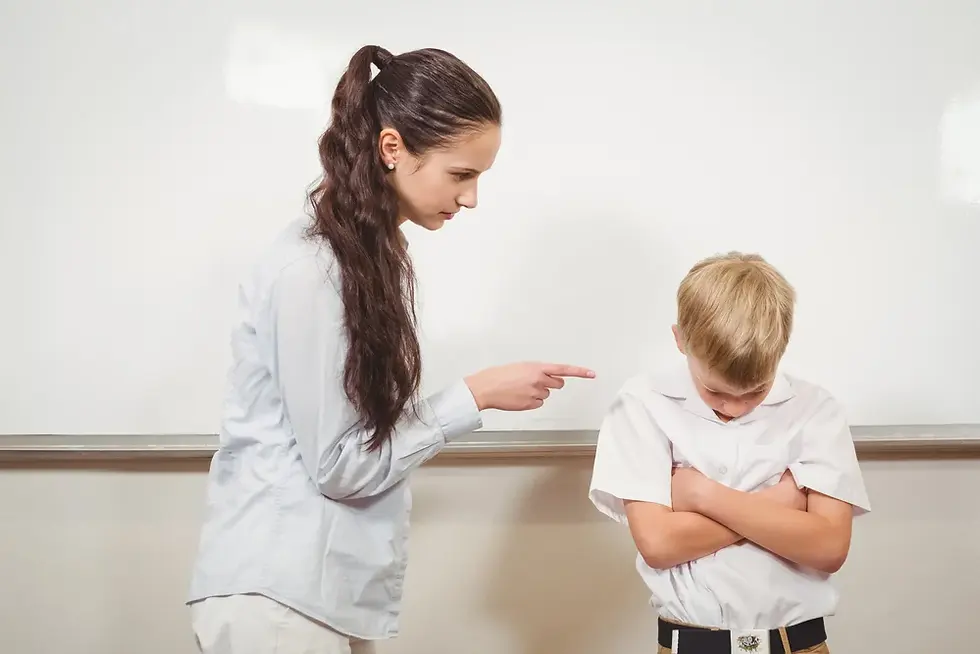 Woman pointing at a boy with crossed arms in front of a whiteboard. The boy appears upset, while the woman seems stern. Classroom setting.