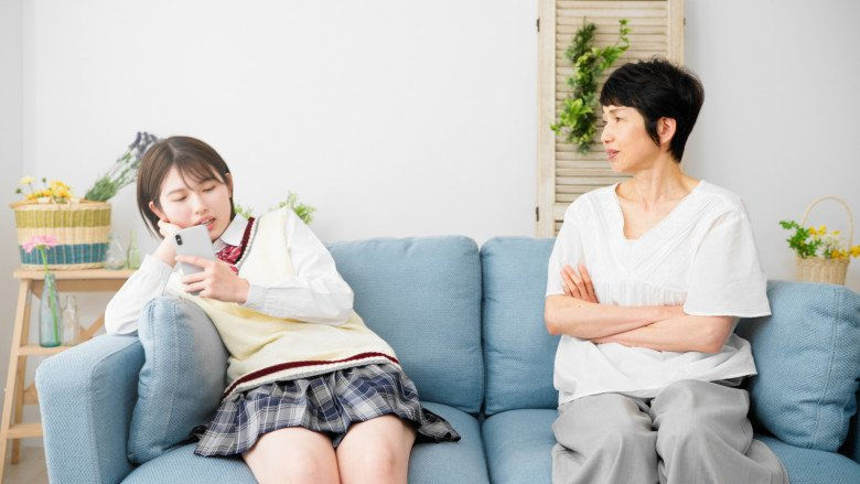 Thiếu nữ xem điện thoại trên sofa xanh, bên cạnh người phụ nữ ngồi khoanh tay, nhìn sang chỗ khác trong phòng khách sáng.