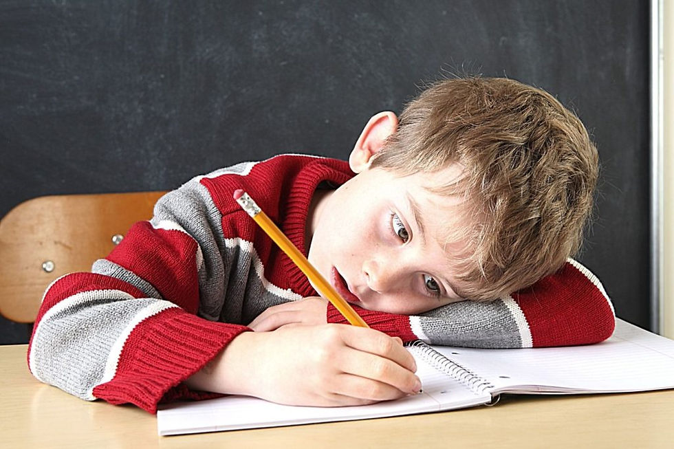 Cậu bé mặc áo len đỏ và xám chống cằm trên bàn học, trông mệt mỏi khi viết vào vở, thể hiện khó khăn tập trung ở trẻ mắc ADHD