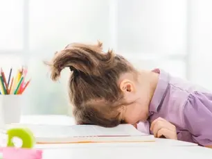 Bé gái ADHD, mặc áo sơ mi tím, đầu tựa vào cuốn sách mở bên chiếc bàn trắng, xung quanh là những cây bút chì đủ màu. Tâm trạng có vẻ mệt mỏi hoặc bực bội.