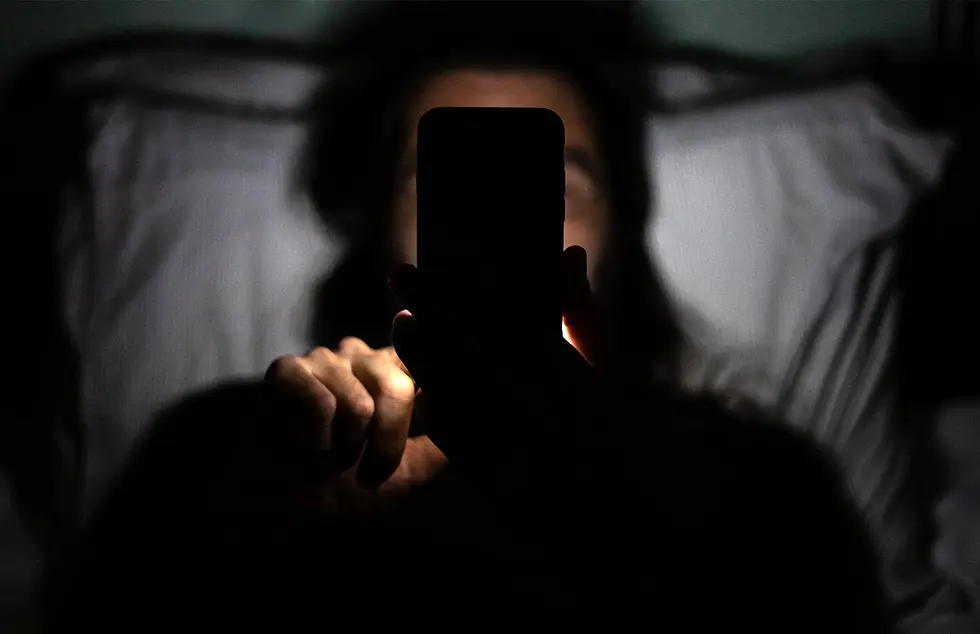 Một người nằm trên giường trong phòng tối, khuôn mặt chỉ được chiếu bởi ánh sáng điện thoại, thể hiện thói quen dùng màn hình ban đêm gây quá tải tinh thần.