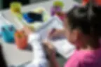Young girl coloring a worksheet with crayons at a table. Various colored cups hold crayons. Bright setting creates a playful mood.