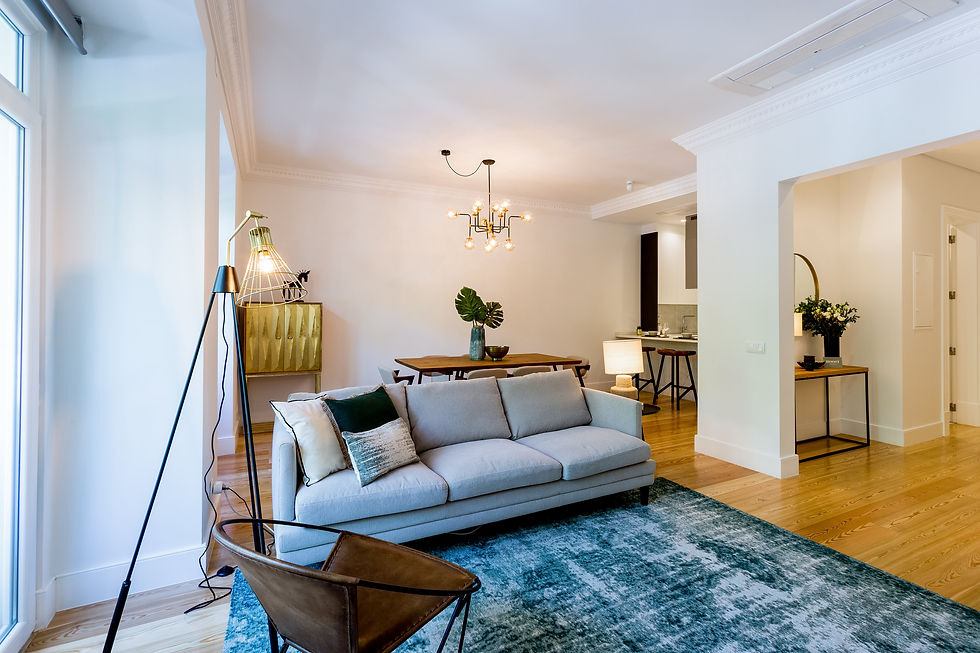 Sala grande aberta para a cozinha. Decoração em tons de ver de azul com sofá grande beje, três almofdas, tapete grande verde, cadeirão em cabedal e pés de metal preto.Candeeiro de pé preto e dourado. ao fundo da sala está um móvel em talha dourada, mesa de jantar com tampo em madeira e pés em vidro. Seis cadeiras bejes, Candeeiro de tecto grande com estilo um pouco industrial, com casquilho das lampadas em dourado