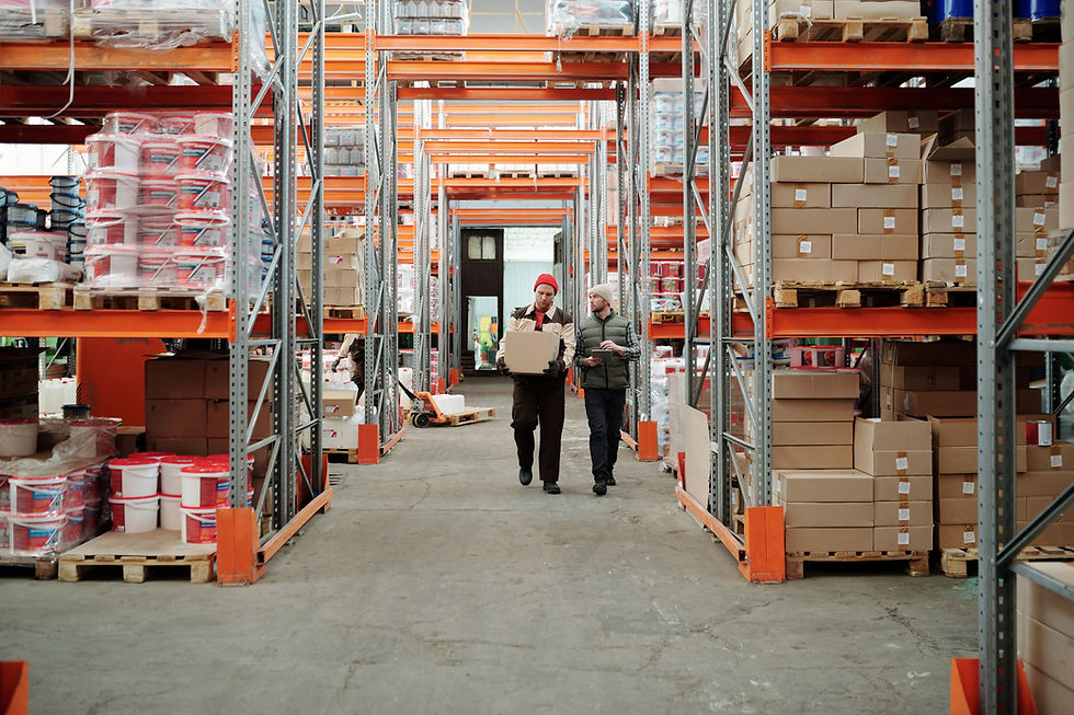 Two men walk through a warehouse aisle, one carrying a box and the other a tablet, between tall shelves of boxes and paint buckets.