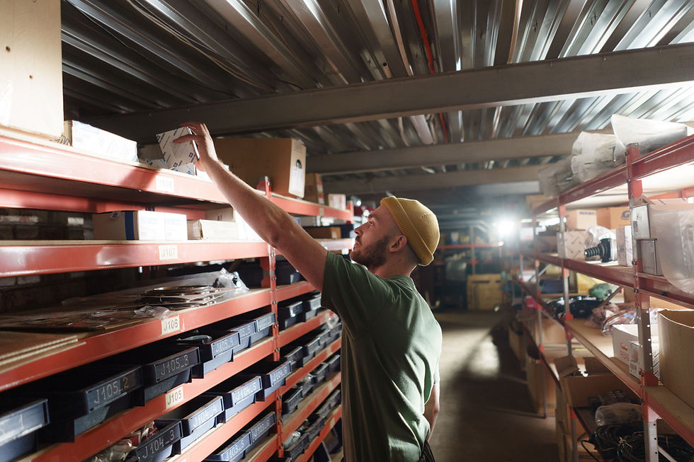 Warehouse worker doing manual picking. He is in yellow beanie reaches for a box on warehouse shelves lined with labeled bins in warm light.