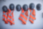 Orange safety vests with reflective stripes and gray hard hats hang on a white wall, creating a neat, organized pattern.