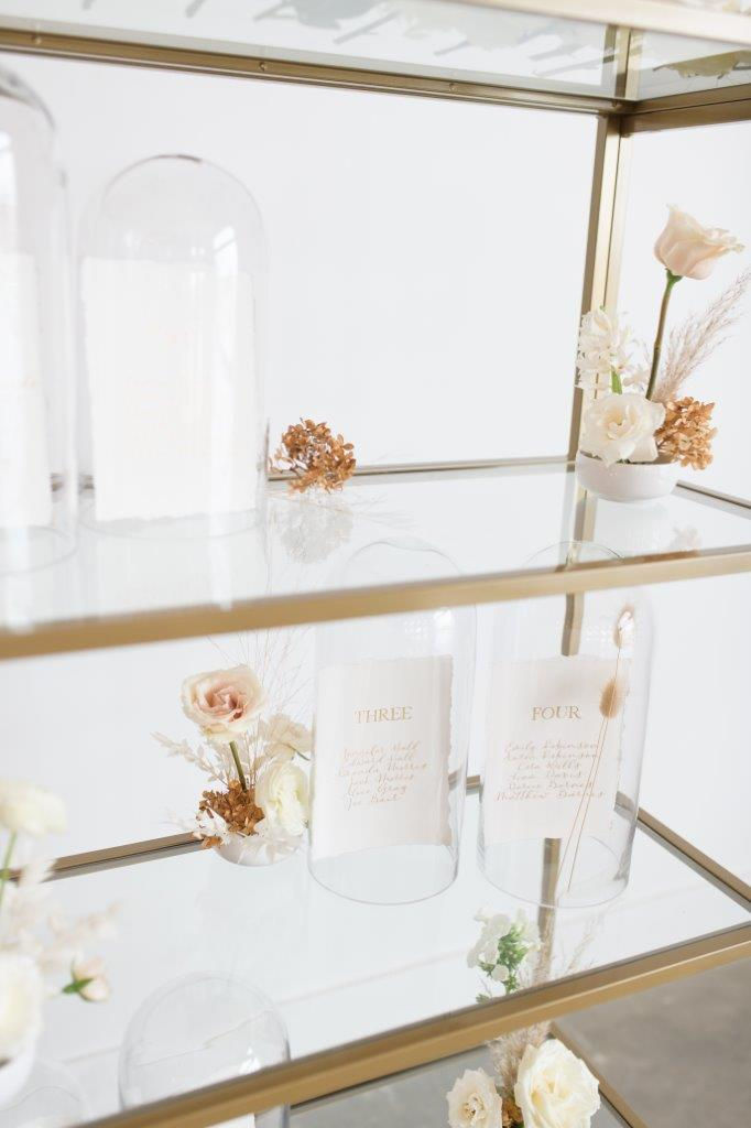 Minimalist wedding seating chart on gold shelves and calligraphy paper inside glass cloche dome with flowers by Studio Bloom