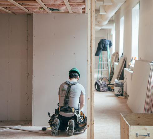 Construction worker on his knees doing renovations to a room. Unfinished drywall and insulation on the ceiling with a ladder in the background with coat on top and tool bag on the ground