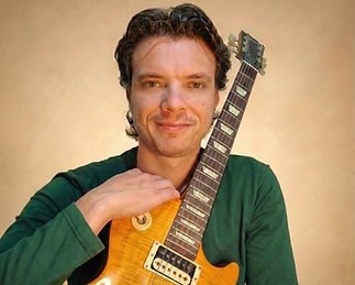 דיוקן של גבר מחייך בחולצה ירוקה, נשען בידו על גיטרה חשמלית צהובה מסוג לס פול, על רקע בהיר במסגרת.
Portrait of a smiling man in a green shirt resting his hand on a yellow Les Paul–style electric guitar, against a light background within a frame.