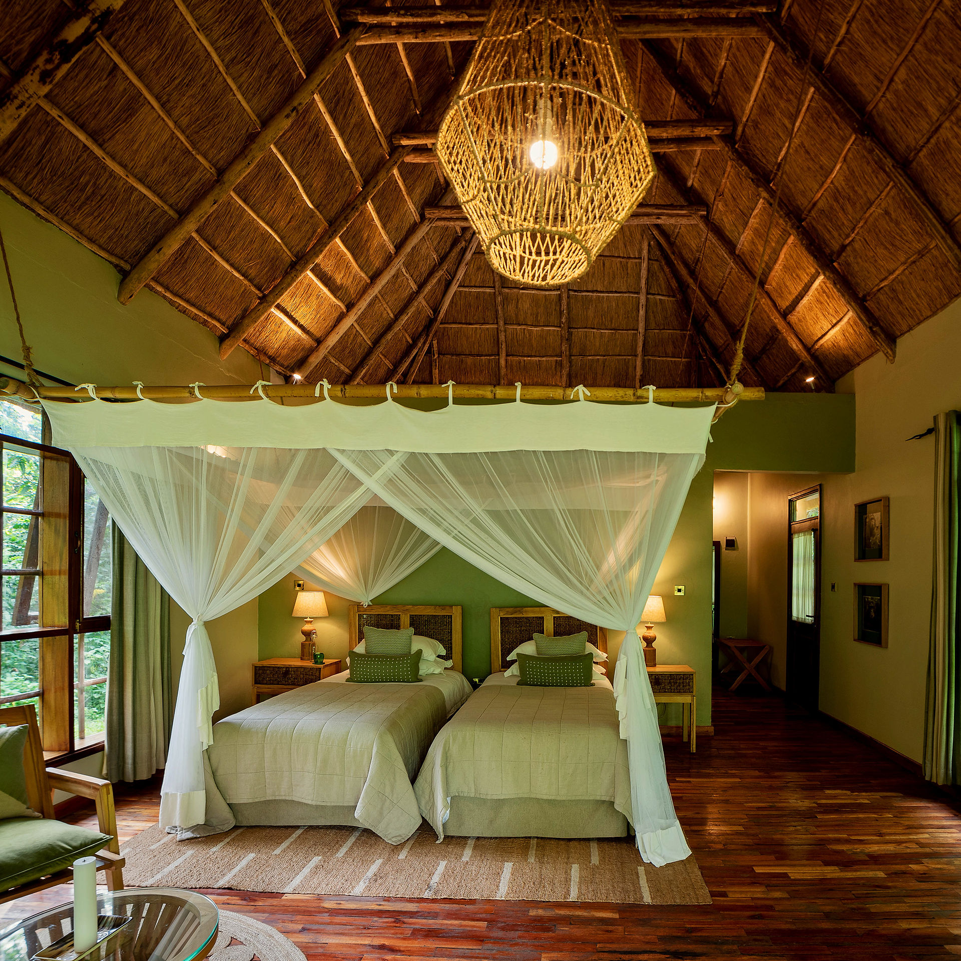 Interior of a twin room at Primate Lodge Kibale, featuring thatched roof, mosquito-netted beds, natural wood finishes, and views into the surrounding rainforest—combining comfort with eco-luxury in Uganda’s jungle.