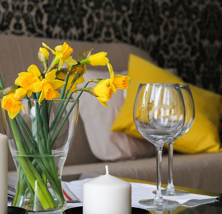 Un living minimalista beige decorado con tips amarillos junto a una vela, dos copas de vino y unas flores amarillas
