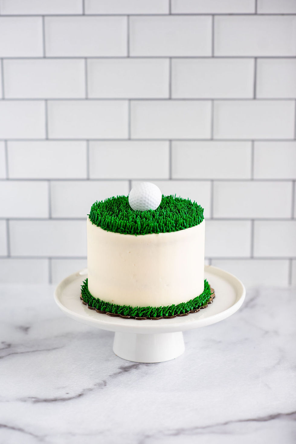 white buttercream smash cake with piped green grass texture and golf ball topper on a white cake stand.