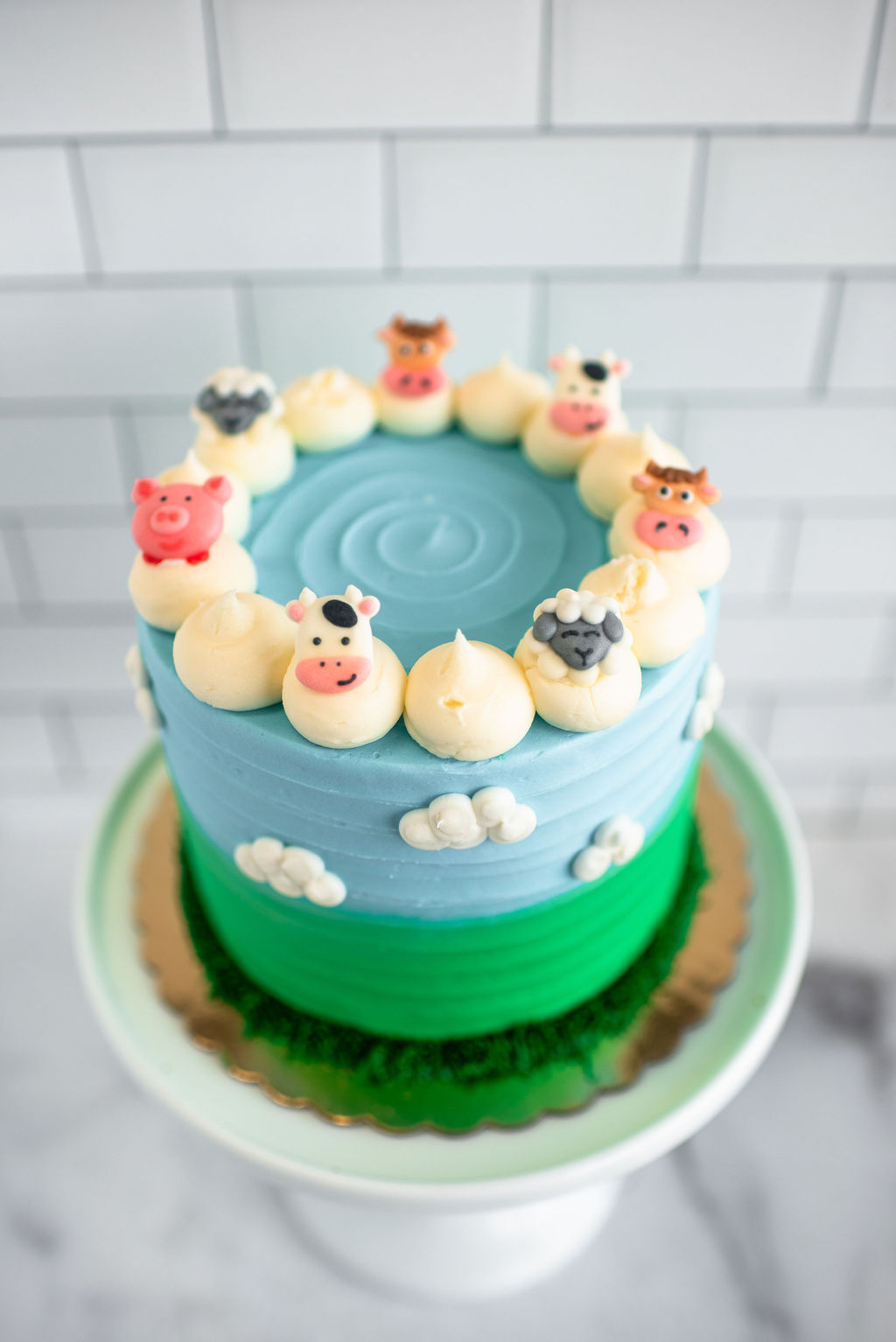 Farm-themed birthday cake decorated with fondant cows, pigs, and sheep, buttercream clouds, and green pasture details on a wh
