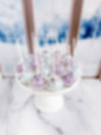 Pastel purple and blue “Frozen Winter” cake pops decorated with sprinkles and displayed on a white cake stand near a snowy wi