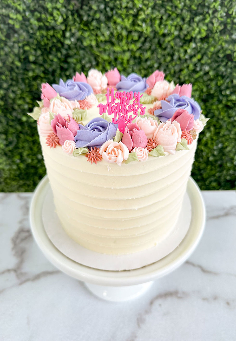 Mother’s Day cake with smooth white buttercream frosting, decorated with pink and purple buttercream flowers and a “Happy Mot