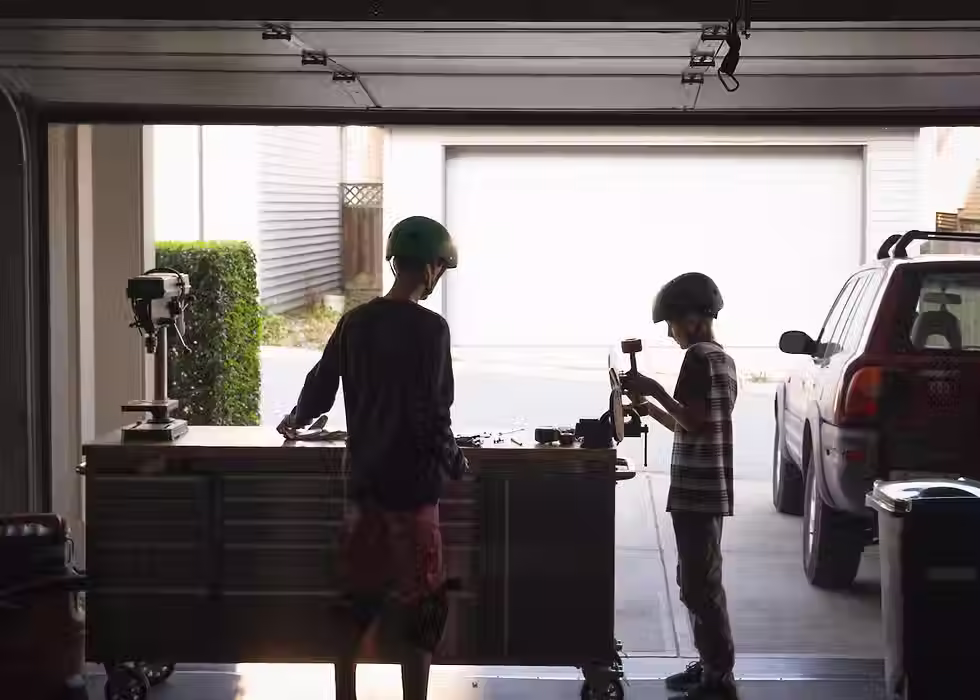 Deux enfants bricolent dans un garage ouvert, casques sur la tête, concentrés sur leur projet.