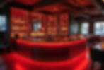 Modern bar with red lighting, two people behind the counter, shelves filled with bottles, and cozy seating. Mood is vibrant and welcoming.