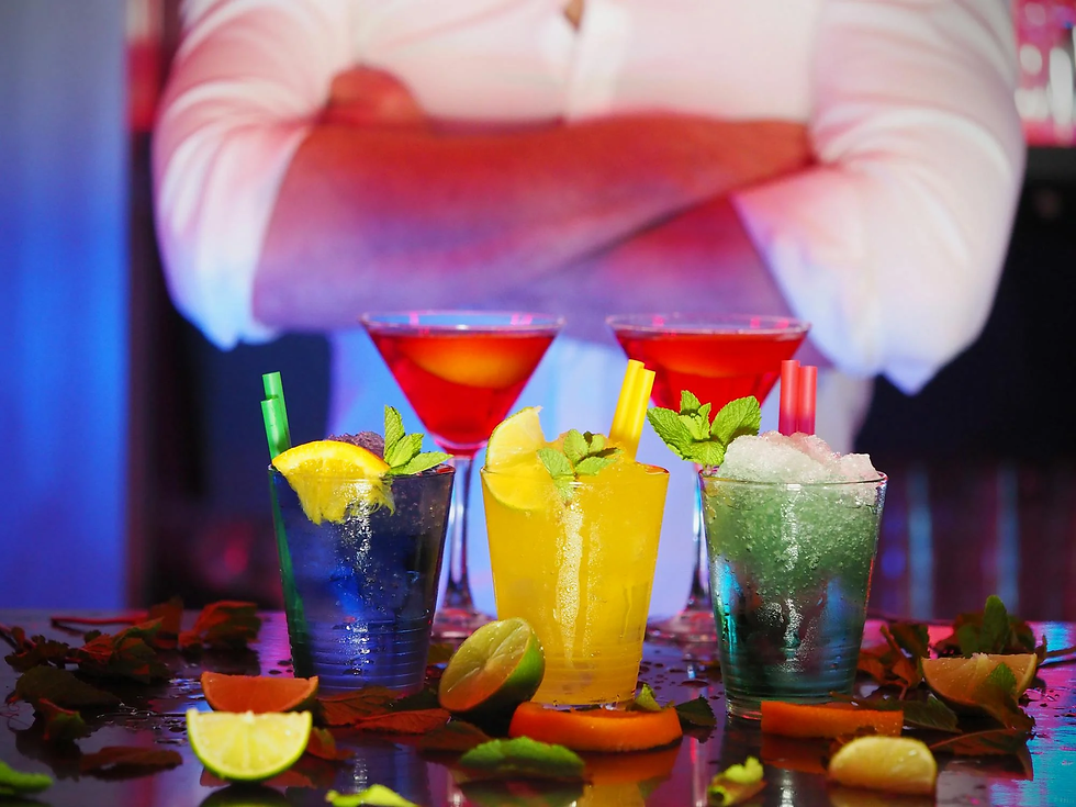 Colorful cocktails garnished with fruit and mint on a bar counter. A person in a white shirt stands in the background, arms crossed.