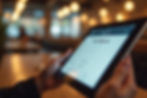 Hands hold a tablet displaying a "Guest Sign up" form. Blurred warm lights and wooden tables create a cozy cafe atmosphere.