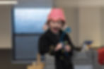 Child looks down, open-mouthed, wearing dress-up hat and holding blue plastic tile