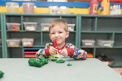 Art room, child smiles at camera, holding play-dough scissors with play-dough on table in front