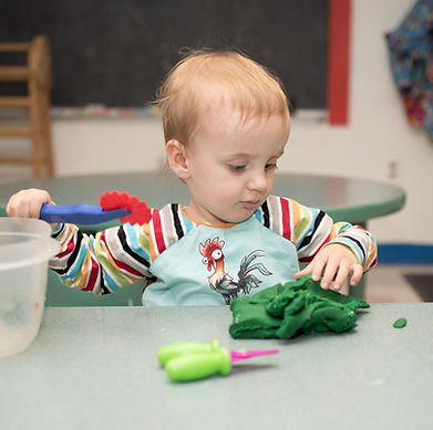 Child in PTLL art room, at table looking at play-dough, a cutting tool in one hand raised above it