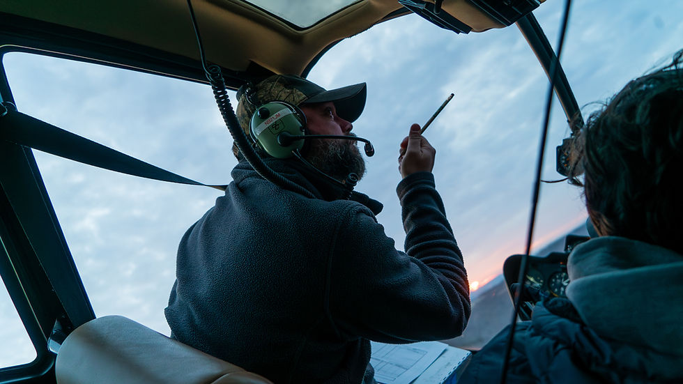 Pilot in a headset talks using a headset in a helicopter cockpit. Blue sky in background, with sun setting. Calm, focused atmosphere.